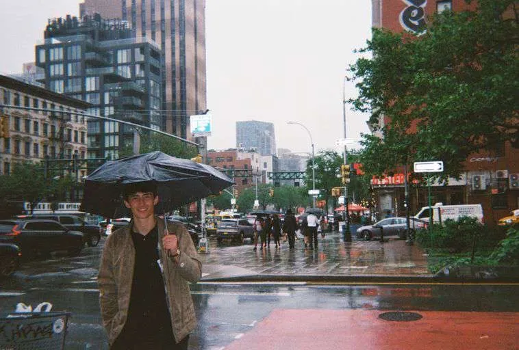 Jackson holding a black umbrella on a rainy New York street corner, wet pavement and traffic behind him