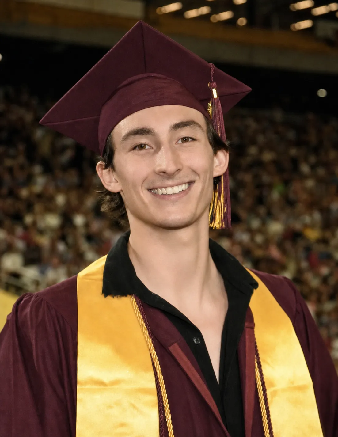 Jackson in a maroon graduation cap and gown with a gold stole and honour cords, smiling in a packed arena